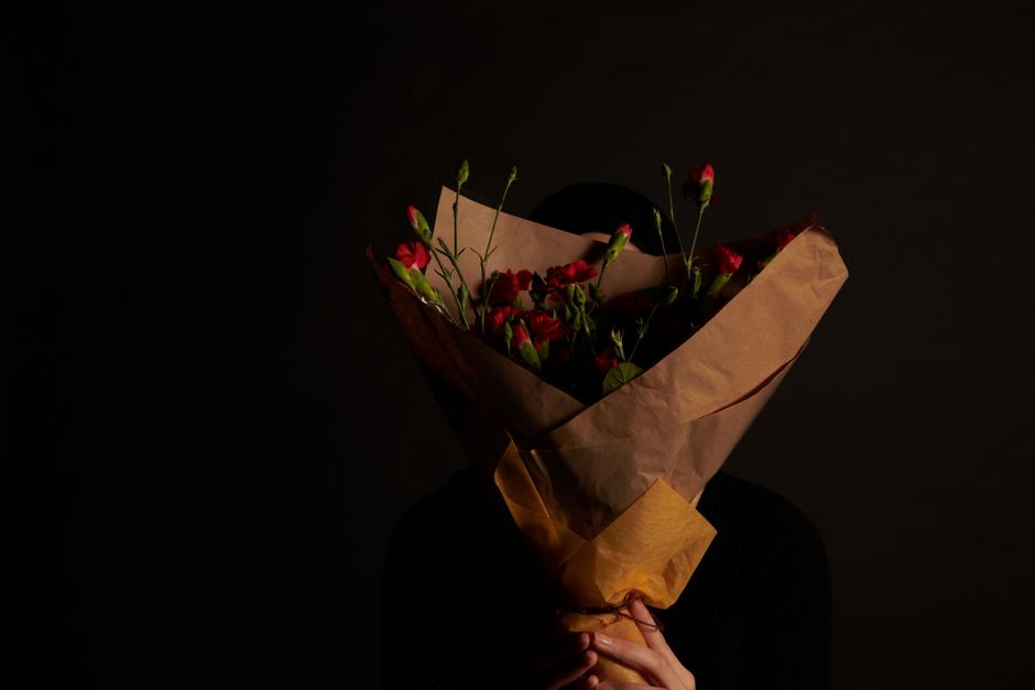 A person holding a bouquet of fresh red and pink flowers wrapped in brown kraft paper with a yellow paper band, set against a dark background. The bouquet includes vibrant, healthy petals with visible