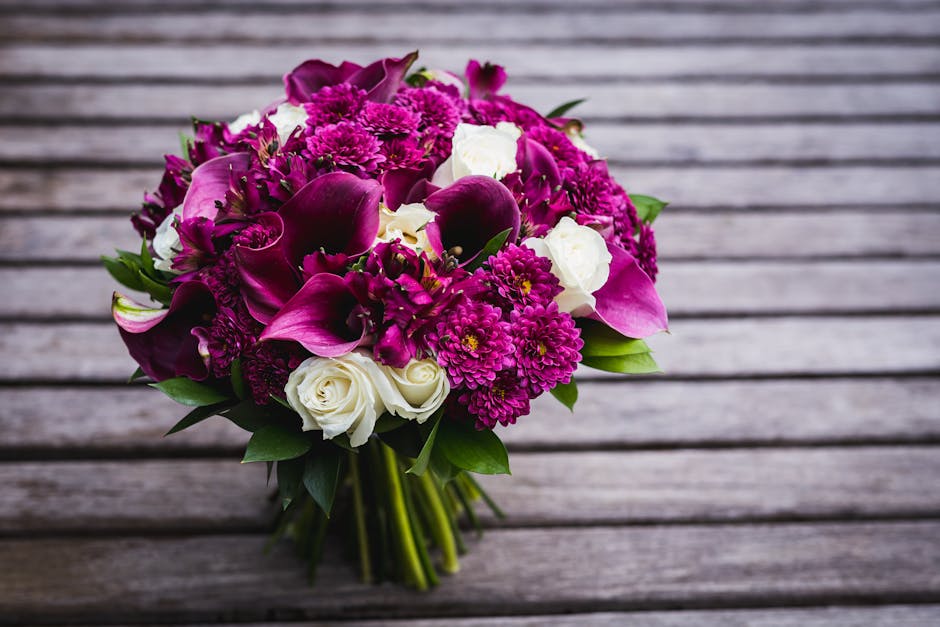 A vibrant bouquet of fresh flowers arranged with deep magenta calla lilies, pink chrysanthemums, and white roses, accented with lush green foliage. The bouquet is held together with transparent wrappi