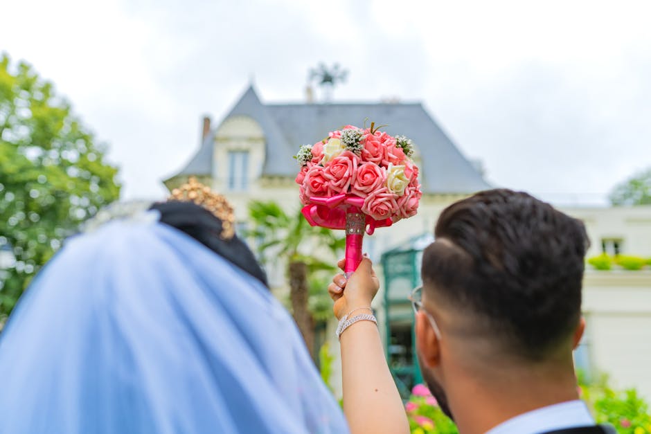 A woman holding a bouquet arrangement of pink and white roses, accented with small white filler flowers, wrapped in vibrant pink paper with a satin ribbon, stands with a man outside a grand historic h