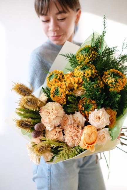 A woman holding a large bouquet of fresh flowers, including pale pink roses, yellow mimosa, and orange carnations, arranged with green foliage and wrapped in light pastel paper. The flowers exhibit vi