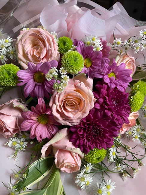 A close-up image of a floral bouquet featuring pale pink roses with tightly curled petals, complemented by vibrant purple daisies with bright yellow centers, and small white daisies with yellow center
