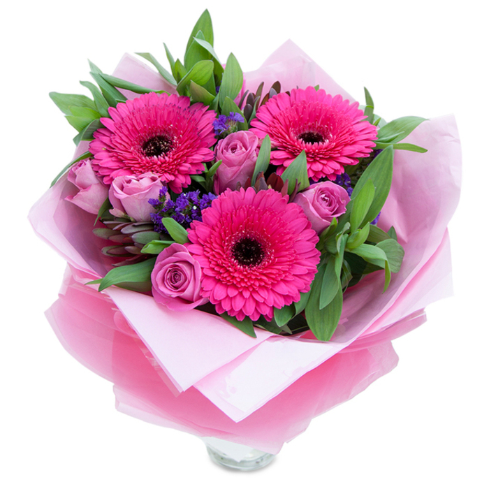 I picture this arrangement as a bright, tender embrace: three deep fuchsia Gerbera daisies open wide like little suns, their dense petals arranged in orderly rays around dark, contrast-rich centres. Nestled beside them, roses in a dusty lavender-pink sit slightly more reserved, their velvety petals arranged in concentric folds that soften the composition and invite touch. Between these major forms are small sprays of purple statice, their delicate, textured clusters bringing an informal, wildflower note that echoes hedgerow charm. The foliage underpins the colour - generous, glossy green leaves that read as salal or eucalyptus, their shapes providing structure and a cooling foil to the pink palette. The bouquet is wrapped with attention to both protection and presentation: a soft inner layer of matte pale pink tissue paper cradles the stems, while a sheer, slightly crinkled fuchsia-pink cellophane overlays it, catching highlights and giving the whole piece a refined, celebratory shimmer. The wrapping has been folded with care to show the top rim of a clear glass vase beneath, signaling freshness and readiness to be displayed on a kitchen island or a mantel. In local light - the gentle afternoon glow that filters through windows in Addington Village near Addington Palace - the colours feel luminous and intimate. The scent is understated: roses imparting a gentle perfume, greens lending a bright, clean note. This bouquet is crafted to make someone feel seen and celebrated, ideal for marking anniversaries, birthdays or simply saying 'you matter' to a neighbour in Addington.